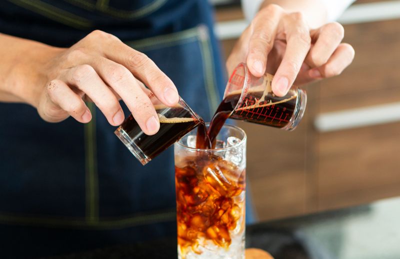 Barista making Ice coffee in a tall glass by pouring brew coffee  over ice on a old rustic wooden table. Young barista pouring cold brew coffee into glass on table. The process of pouring fresh brew coffee into a glass.
