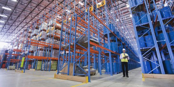 Worker in safety gear inside large warehouse with blue barrels.