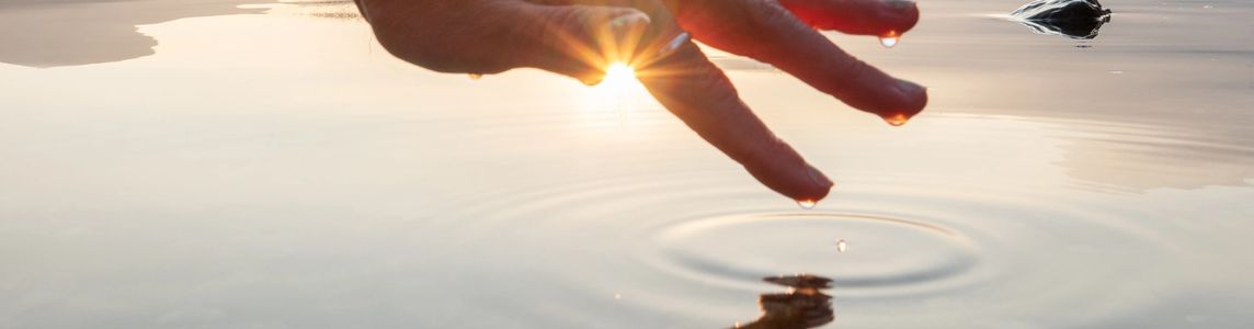 Hand touching water surface with sun shining through fingers at sunset.