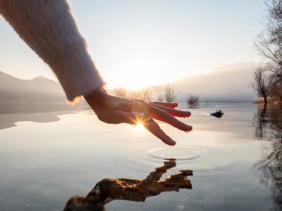 Hand reaching out over calm water during sunset with clear reflection.