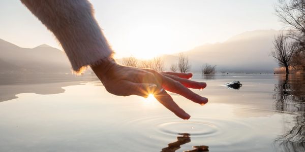 Hand touching calm lake water with sun setting behind mountains.