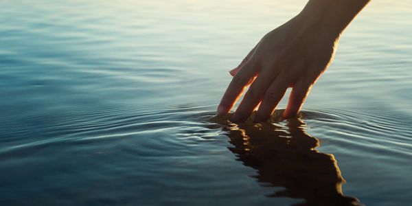 Hand touching calm water surface at sunset.