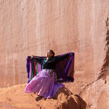 Person in colorful traditional clothing poses on a rock against a desert cliff.