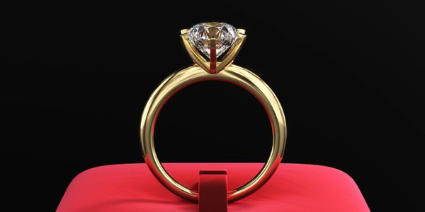 Elegant gold ring with a large diamond on a red cushion against black background.