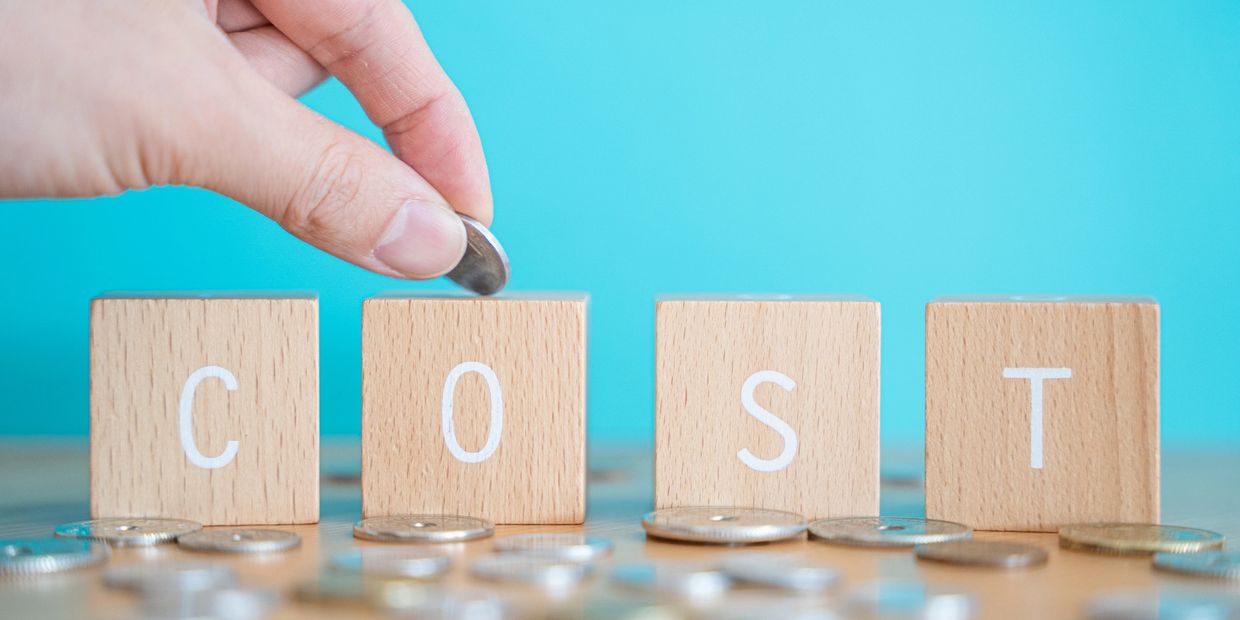 Hand placing a coin on wooden blocks spelling COST with scattered coins.