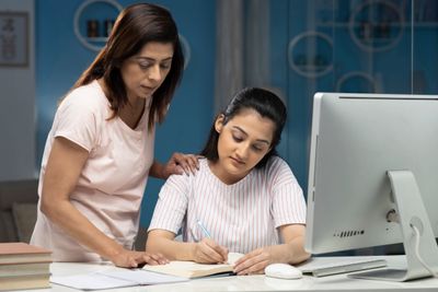 A woman helps a girl with her homework at a desk with a computer.