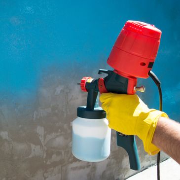Person spraying blue paint on a wall using a paint sprayer with a yellow glove.