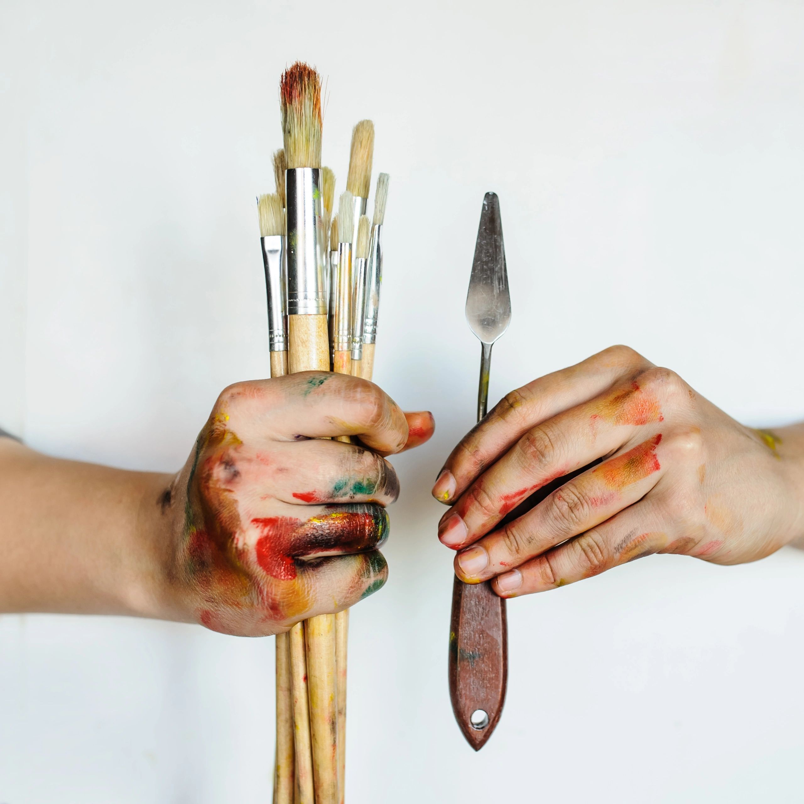 Hands holding paintbrushes and a palette knife with paint stains.
