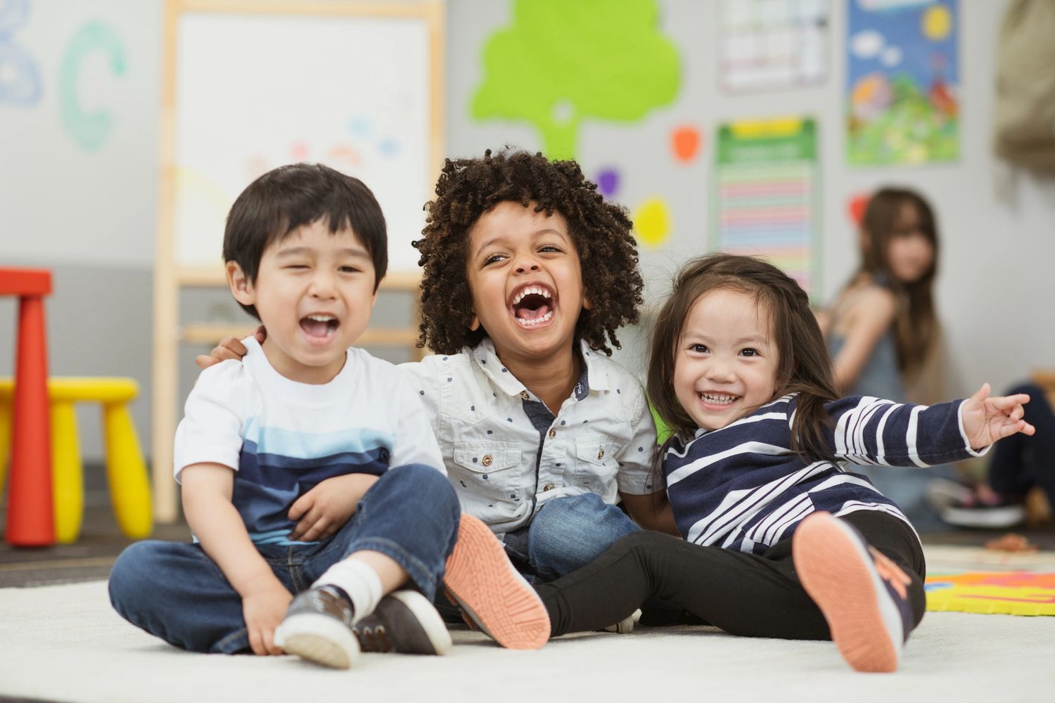 Three joyful children sitting together and laughing in a colorful classroom.