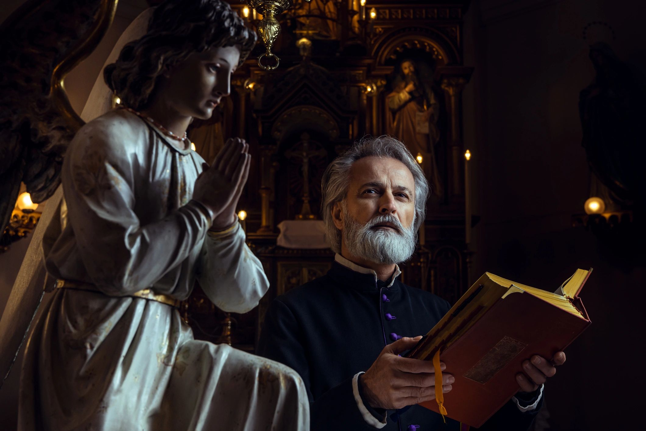 Priest holding a book next to a praying angel statue in a church.