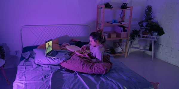 Woman reading and watching laptop on bed with purple lighting.