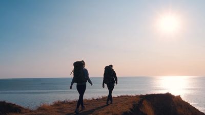 Two hikers walk along a coastal cliff at sunset.