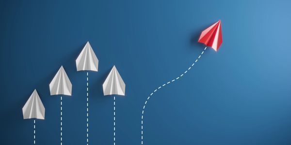 Red paper plane diverges from white planes on blue background, symbolizing innovation and leadership.