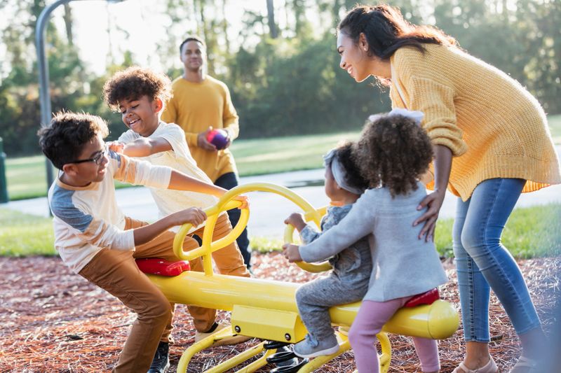 A mixed race family with four children having fun at the playground. The father is African-American and the rest of the family is mixed race African-American and Hispanic. The two girls and two boys are playing together on a seesaw. The parents are in their 30s and the children are 21 months to 10 years old.