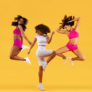 Three women in activewear joyfully jumping against a yellow background.