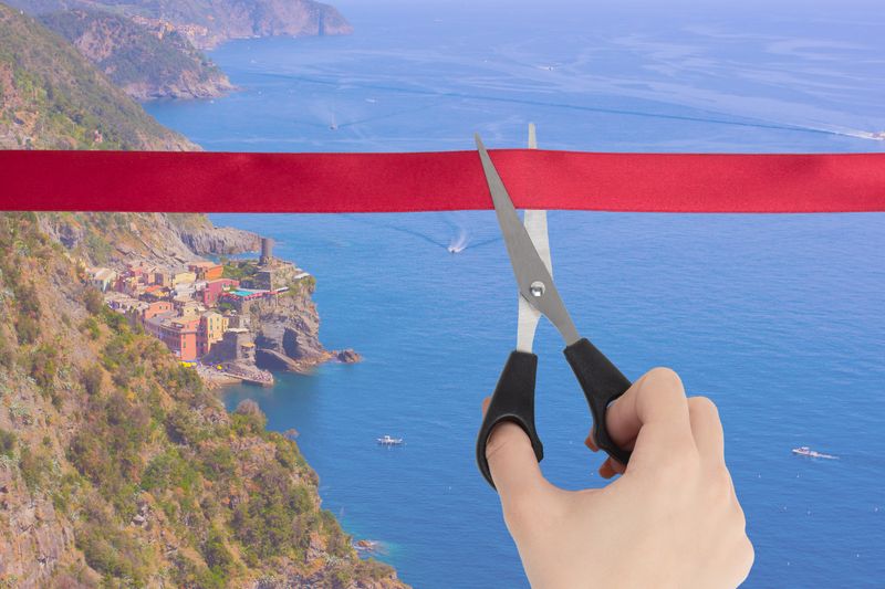 The end of the quarantine of the coronavirus pandemic, the opening of the borders of countries,  the removal of restrictions on air travel, last-minute tours. A hand cuts a red ribbon with scissors overlooking the sea, the coast of the village of Varnaca, Italy