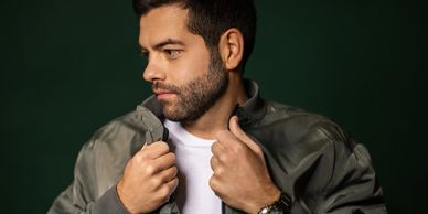 Man with beard adjusting his jacket against a dark green background.
