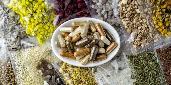 Herbal supplement capsules surrounded by various dried herbs and natural ingredients in plastic bags.