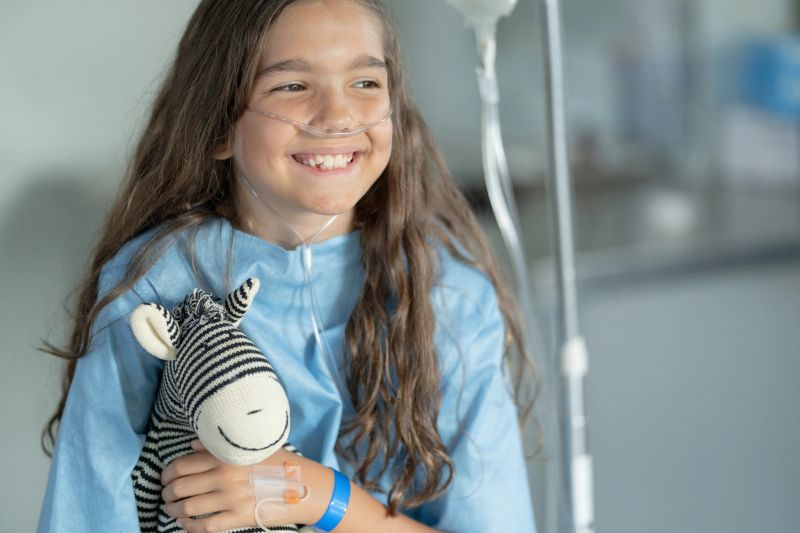 A ten year old girl of Hispanic ethnicity is at the hospital. She has an IV drip and oxygen tube on her nose. She is holding her favourite teddy bear and smiling.