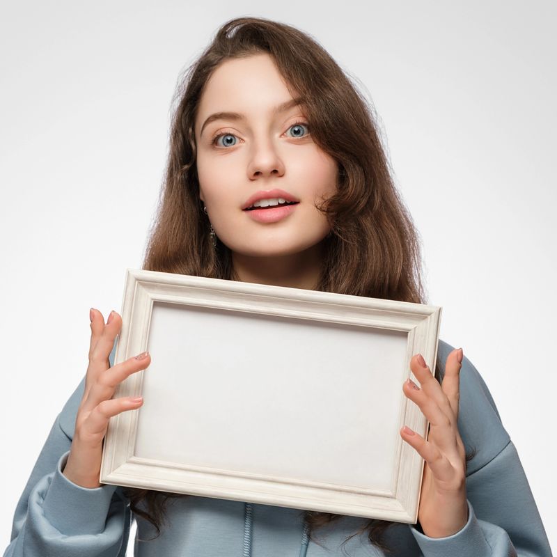 Young woman with an empty picture frame horizontally in her hand on a light blue background. The frame has a space for your text or photo. Light blue hoodie, blue eyes long hair.