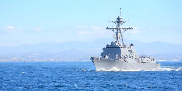 A navy warship sailing on a calm sea with mountains in the background.