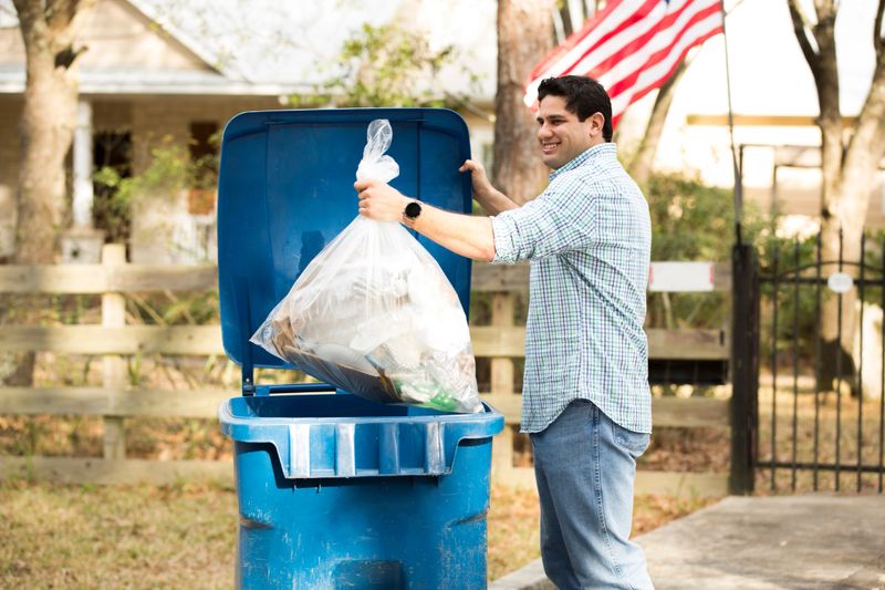 Mid-adult Latin Descent man puts recycle trash in bin.  Bin is located on curb in front of home and is picked up twice a week.  American flies from the fence post.