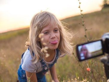 Young girl in denim overalls sticks out her tongue to a camera outdoors.
