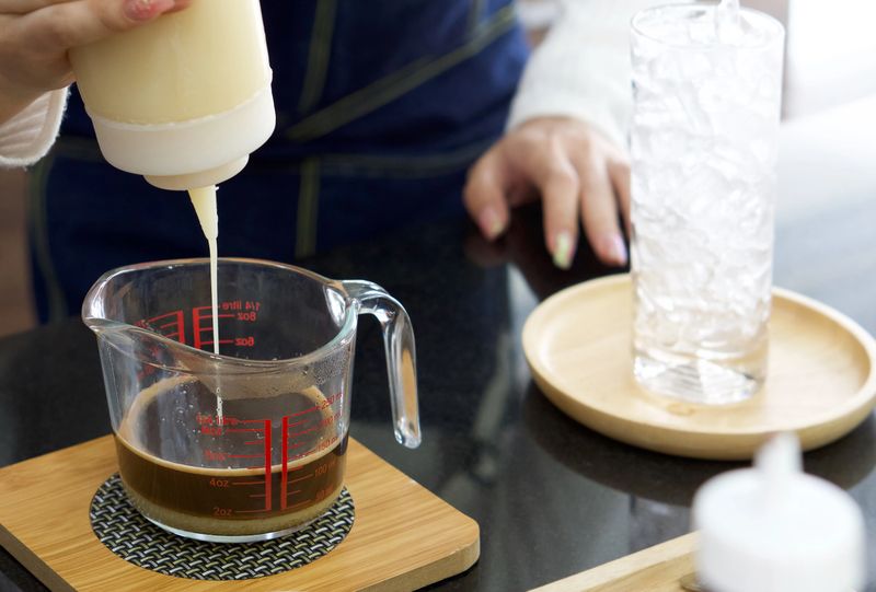 Asian barista squeeze condensed milk from a plastic bottle into a coffee measuring cup. Morning atmosphere in a coffee shop.