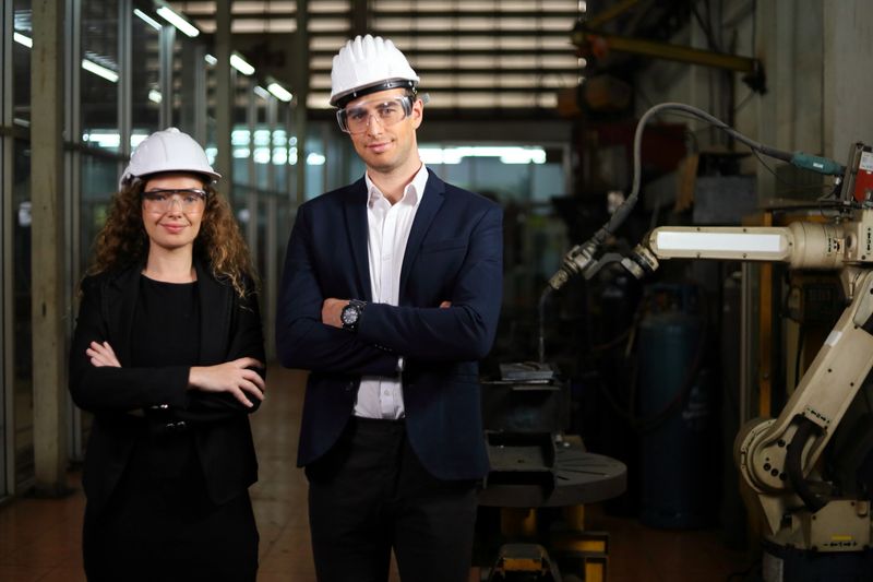 Portrait of two Caucasian engineers standing beside welding robot machine arm inside mechanic factory