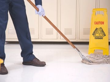 A janitor mops a shiny floor near a yellow caution sign in a locker room.