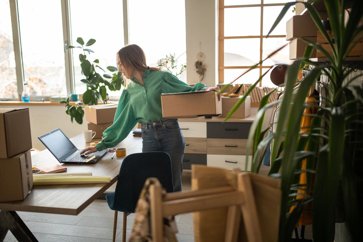 Woman working with laptop and holding a cardboard box in a bright room.
