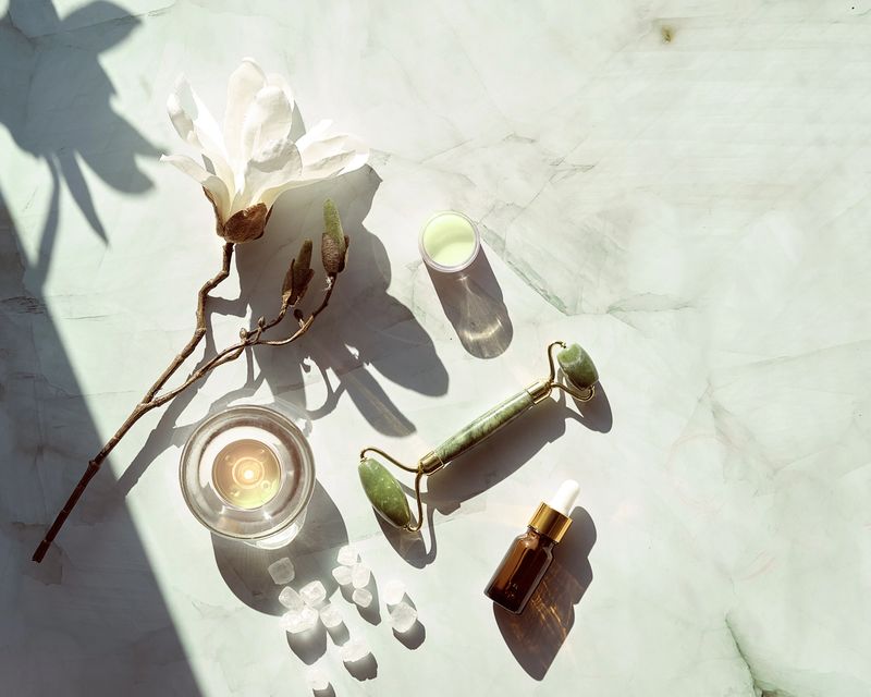 Jade face roller for beauty facial massage therapy, Essential oil bottle. Flat lay on marble table with magnolia flowers. Long shadows and candle.