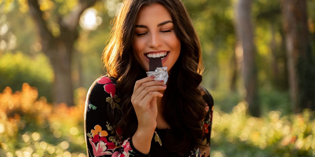 A woman joyfully bites into a chocolate bar outdoors in warm sunlight.