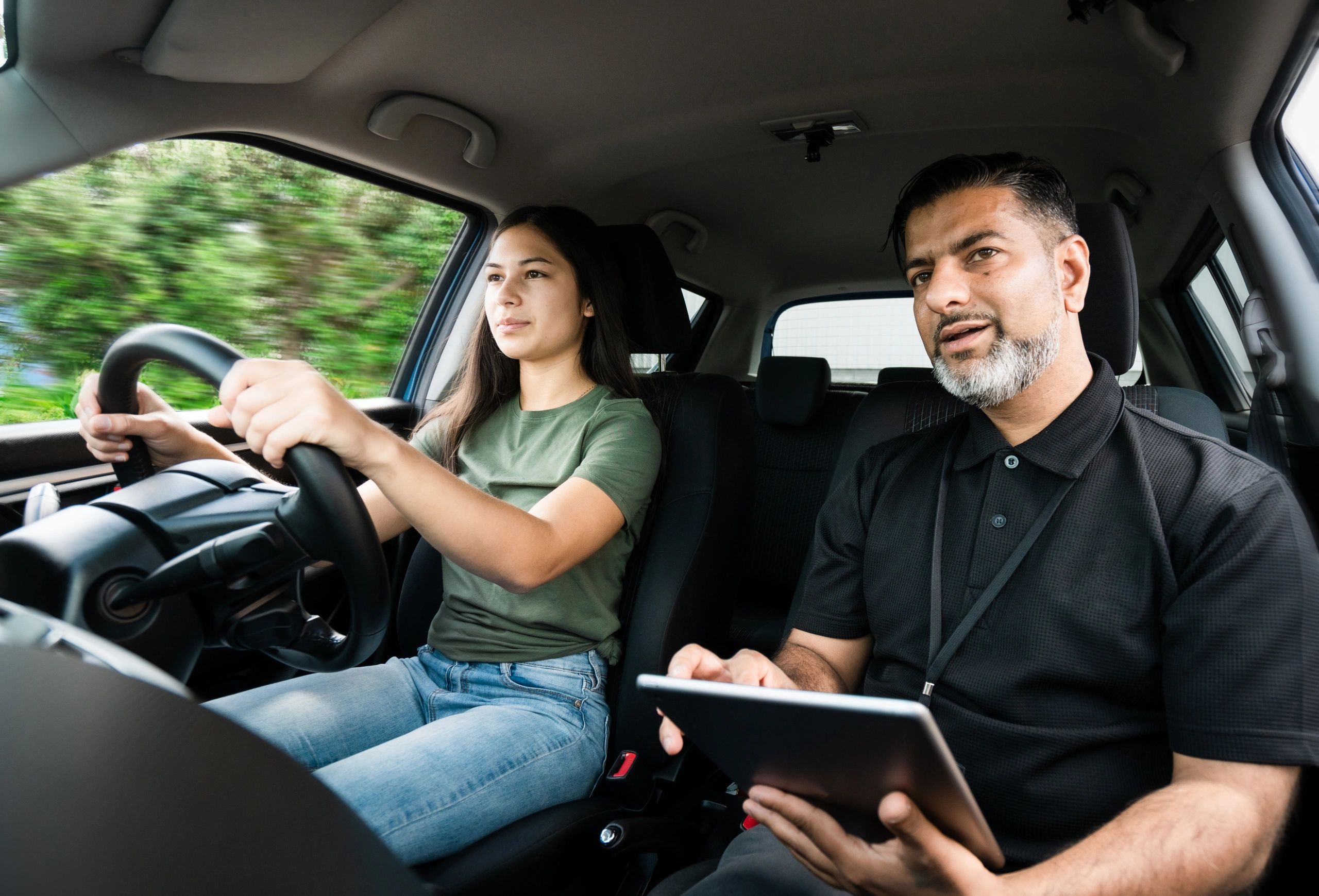 A young woman driving a car with an instructor beside her holding a tablet.