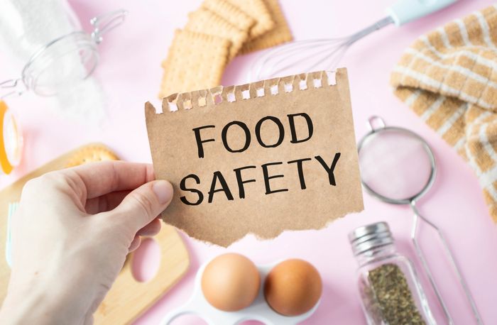 Hand holding a paper with 'FOOD SAFETY' written, surrounded by kitchen items.