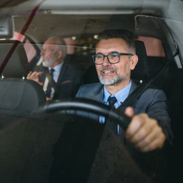 Man in glasses driving a car with an older man in the backseat.
