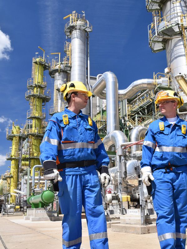 Two industrial workers in blue uniforms and yellow helmets at a refinery plant.