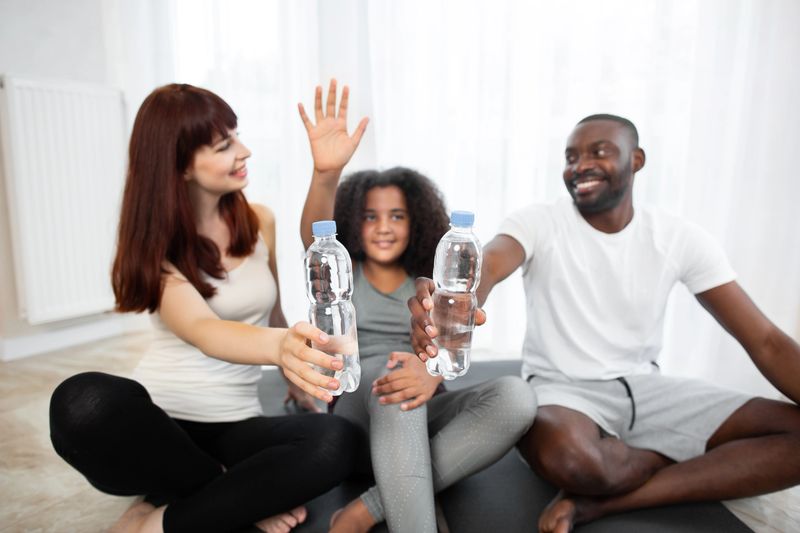 Training at home. Full length blurred view of beautiful teenage girl and her multiethnic parents in sports clothing training yoga on the mat at home and holding bottles with water. Focus on bottles