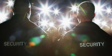 Two security guards face a crowd of photographers with bright camera flashes.