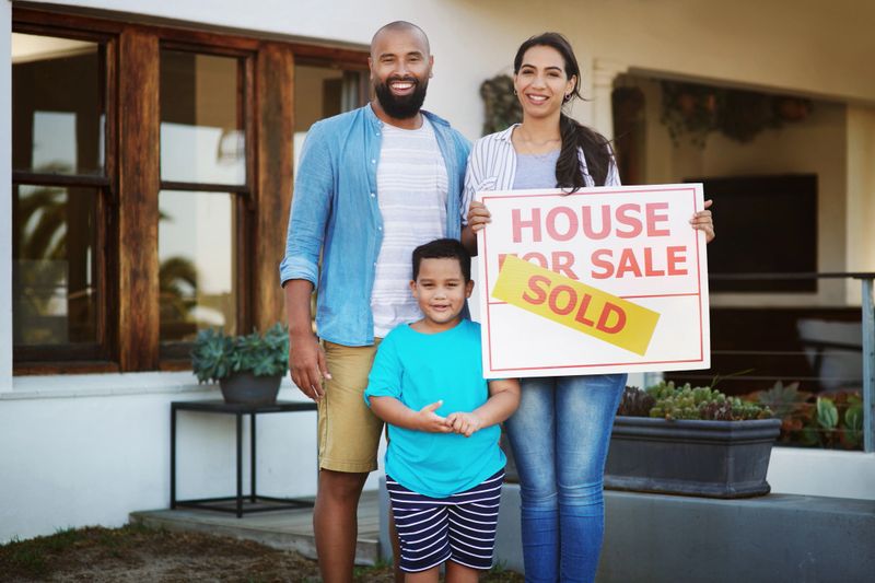 Shot of a young family of three standing beside a sold sign in front of their new home