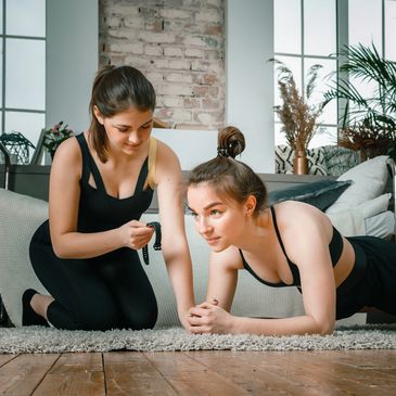 Two women exercising indoors, one doing a plank while the other coaches.