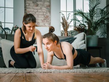 Two women exercising indoors, one doing a plank while the other coaches.