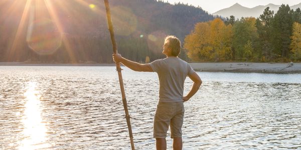 A man stands on a tree stump by a lake, holding a long stick, gazing at the sunlit mountains.