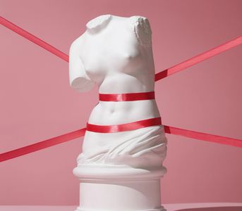 Escultura clásica envuelta con cintas rojas sobre fondo rosa.