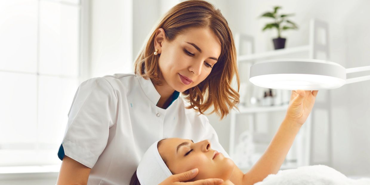 A beautician examines a client's face under a bright lamp in a spa.