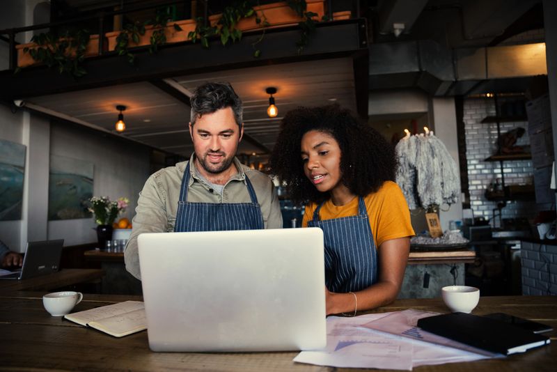 Caucasian manager and ethnic waitress deciding on construction work of cafe sitting with laptop and paper work in trendy coffee shop. High quality photo