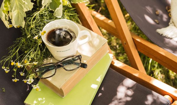 A cup of coffee, glasses, and books on a chair outdoors.