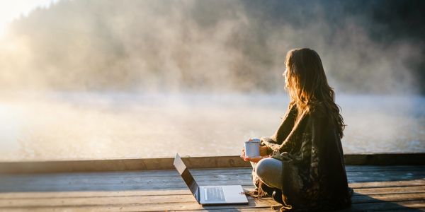 Woman with a cup beside laptop on dock by misty lake.