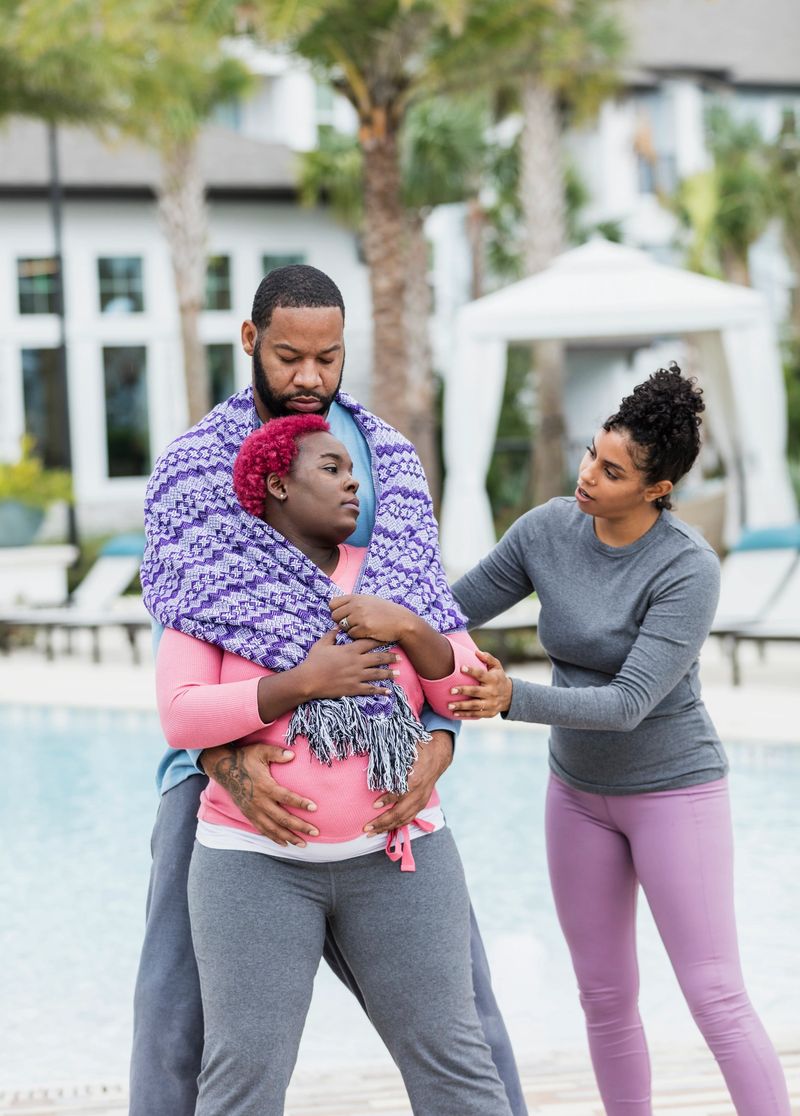 A pregnant African-American couple standing outdoors, by a swimming pool, getting advice from their doula or birth support coach. The expectant mother, who has pink hair and is wearing a pink shirt, and her husband are both in their 30s.