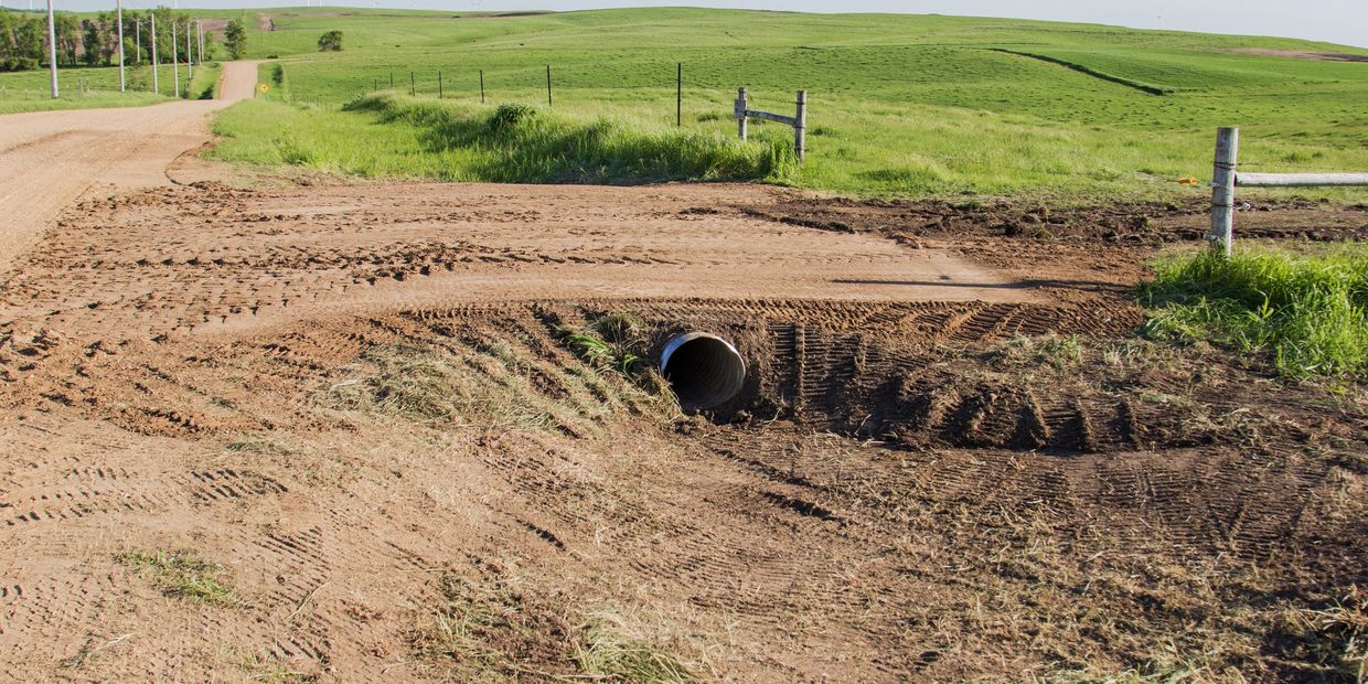 Dirt road with drainage pipe and green fields.
